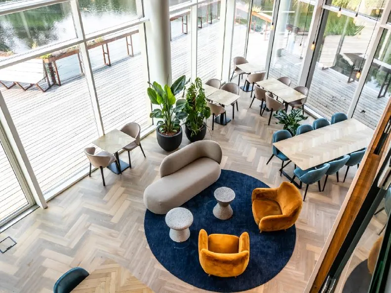 Moderne lounge- en werkruimte aan het John M. Keynesplein met grote ramen, kleurrijke meubels en uitzicht op een houten terras aan het water.